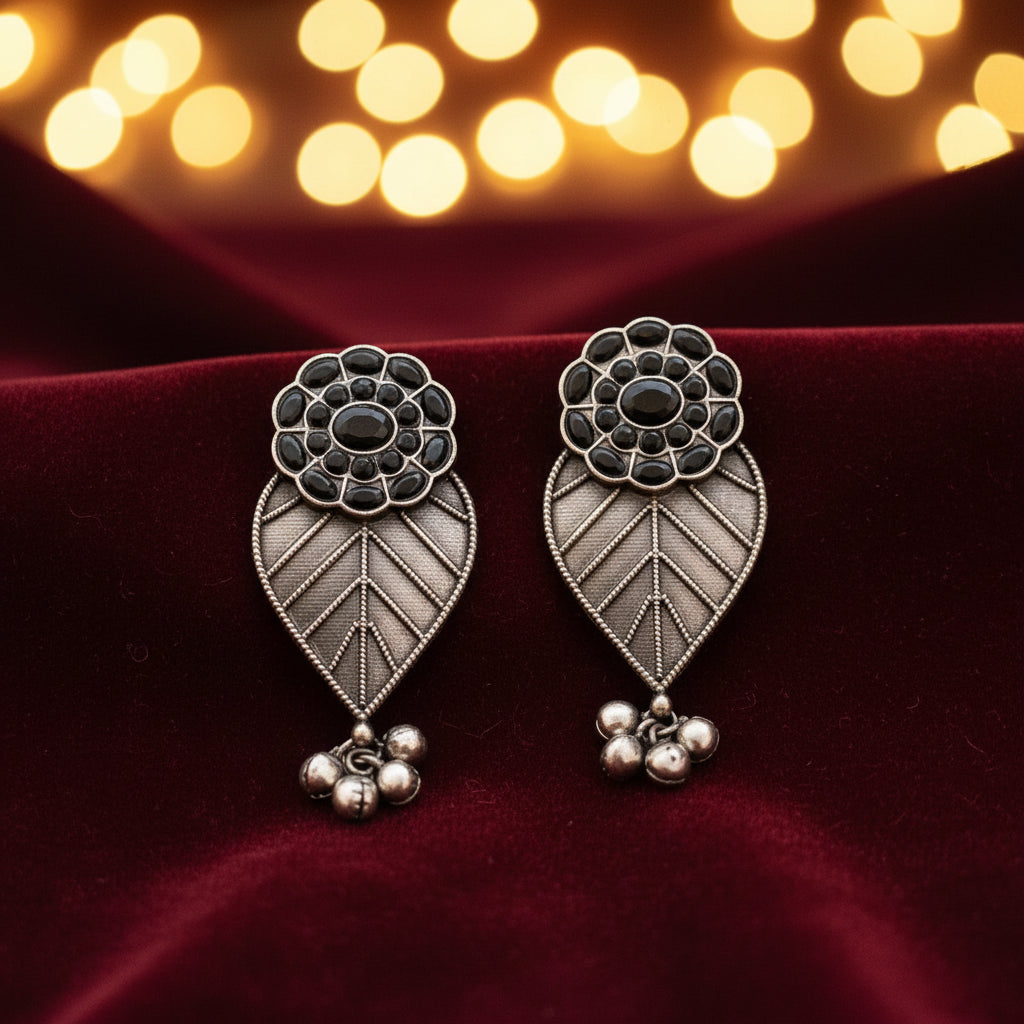 A pair of silver brass earrings designed with leaf patterns and adorned with black stones, displayed against a natural background with a leaf.